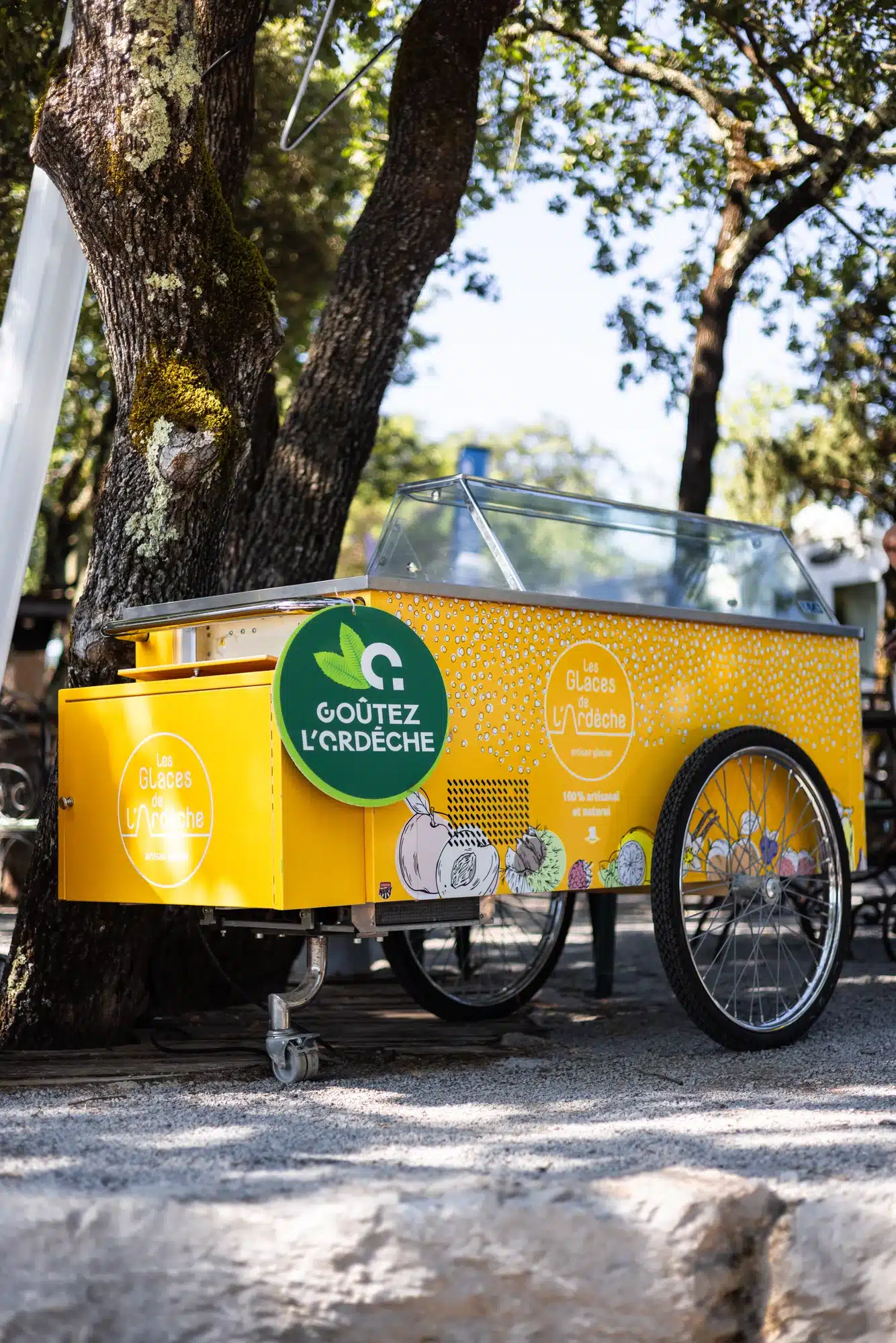 Un chariot jaune vif de glaces artisanales « Les Glaces de l'Ardèche » près d'arbres.