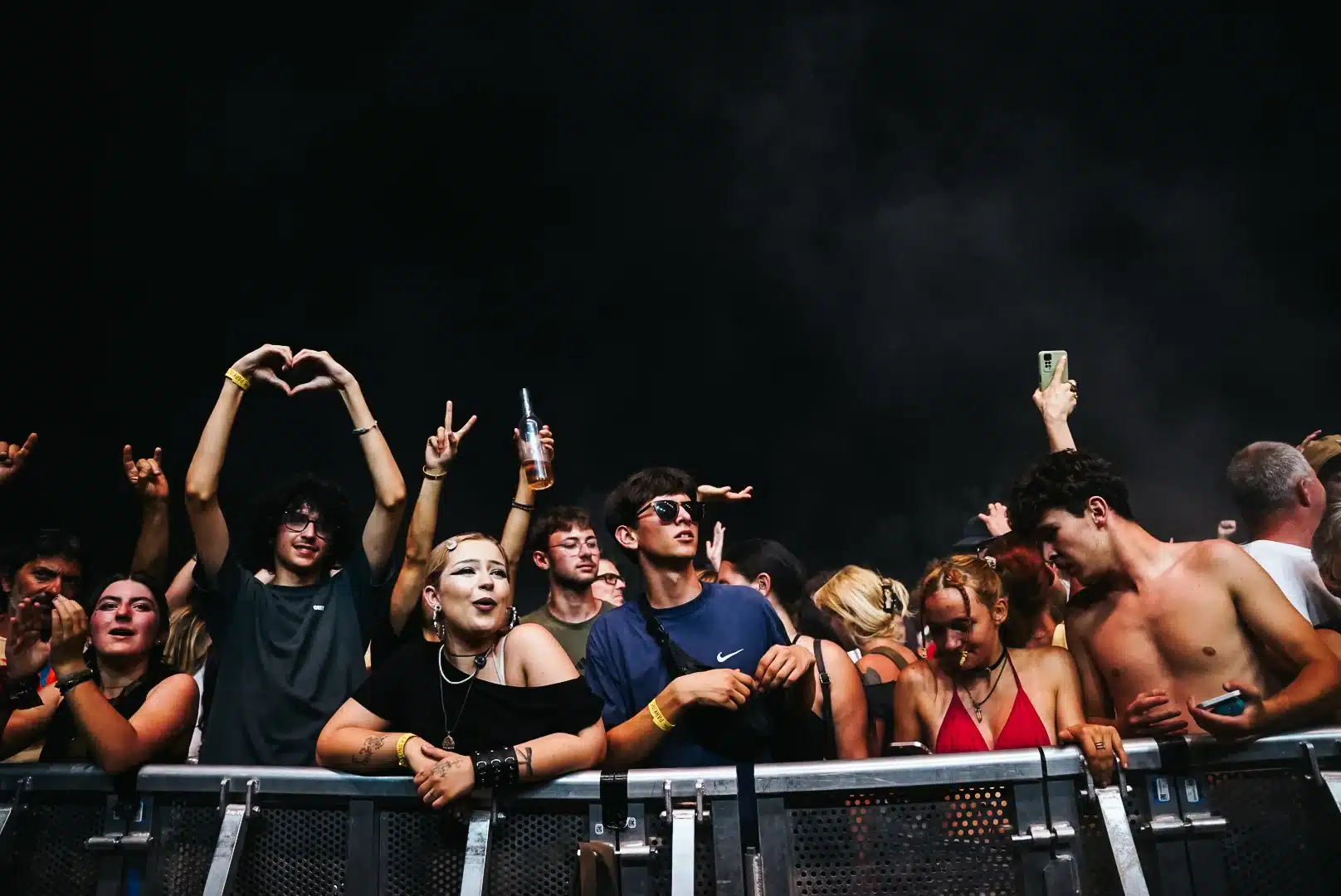 Public d'un festival, jeunes adultes derrière une barrière, mains levées, ambiance festive.